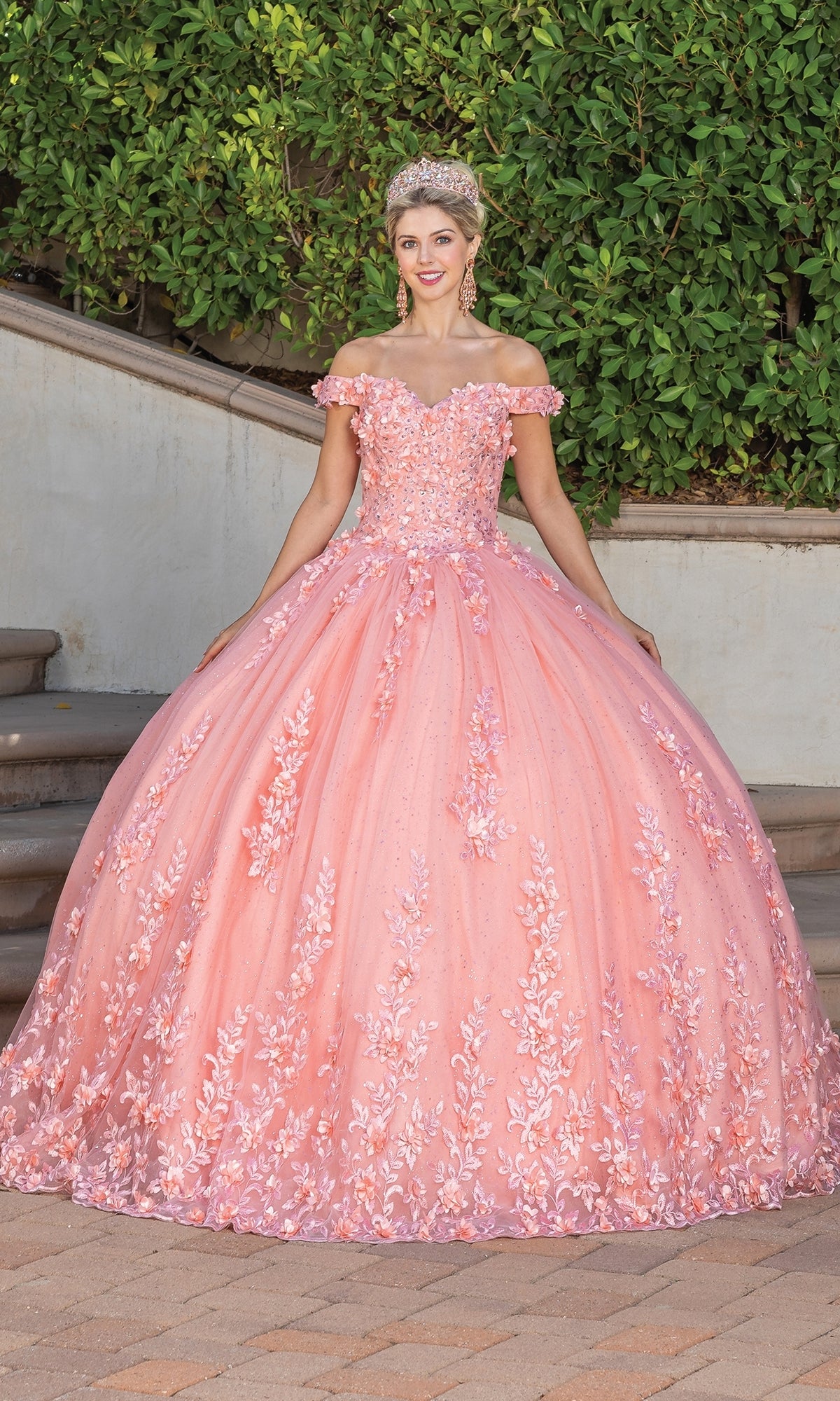 A young woman stands outdoors in the Quinceanera Dress 1727 by Dancing Queen, featuring a coral pink ball gown with floral embellishments, a sweetheart neckline, and a tiara.